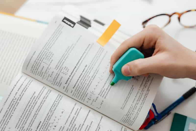 A person marking a line of printed text with a blue highlighter pen, other pages and a notebook on the desk beside them
