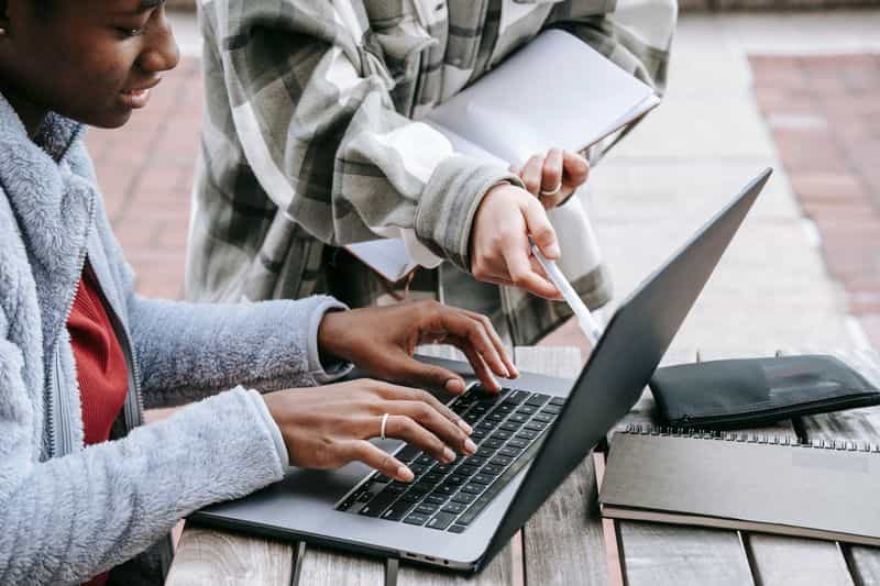 Two students at a laptop on a campus bench, one pointing at the screen while the other types, notebooks beside them