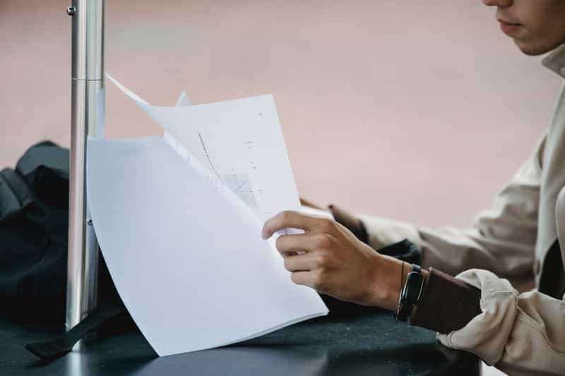 A student reading a printed document at a desk beside an open laptop