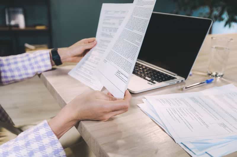 Hands holding two printed pages side by side at a desk beside an open laptop, with more papers and a pen on the table