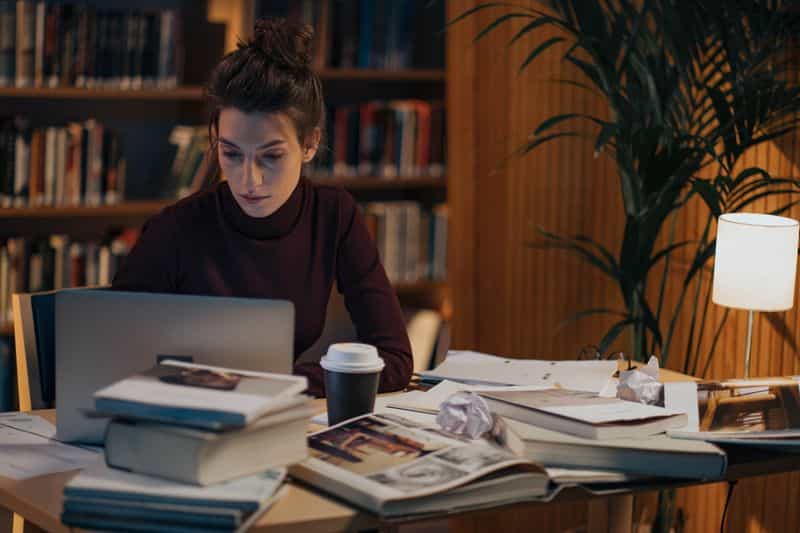 A student working at a laptop in a library, desk covered in stacked books, open journals and a coffee cup