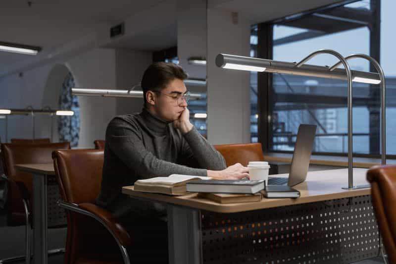 A student in glasses working at a laptop at a library desk, with a stack of books and a coffee cup beside him