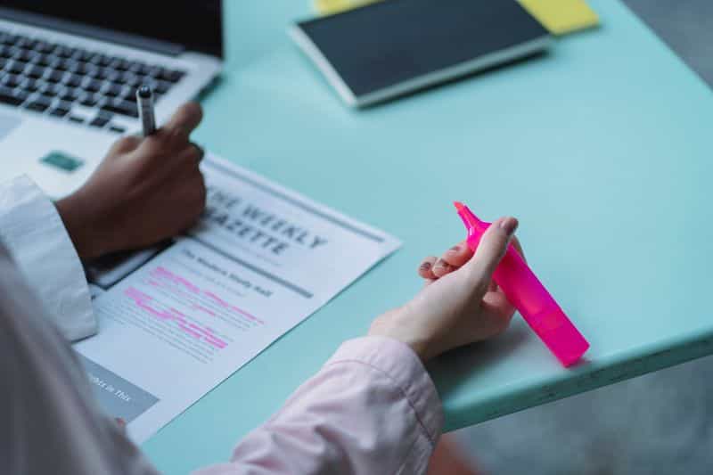 Two people at a desk beside an open laptop, one holding a highlighter over a marked-up printed page
