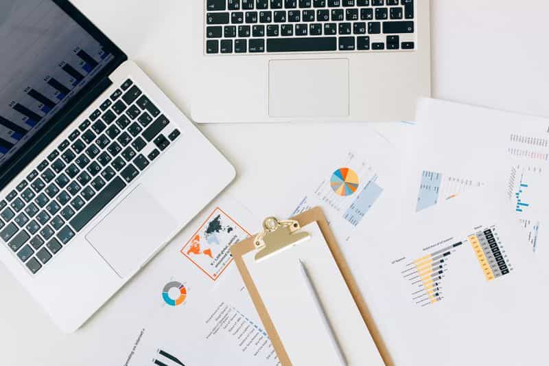 Printed pages of charts and data spread beside two open laptops on a bright desk