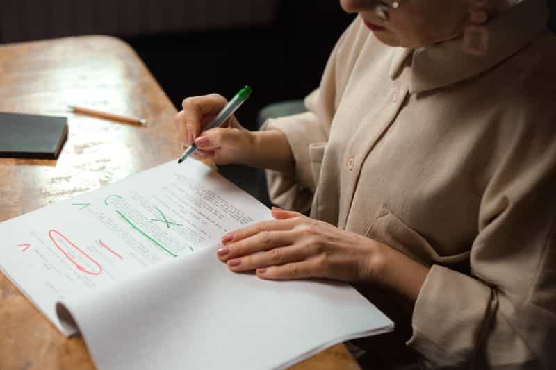 A person at a wooden desk marking up a printed document with a pen, corrections visible in red and green on the page