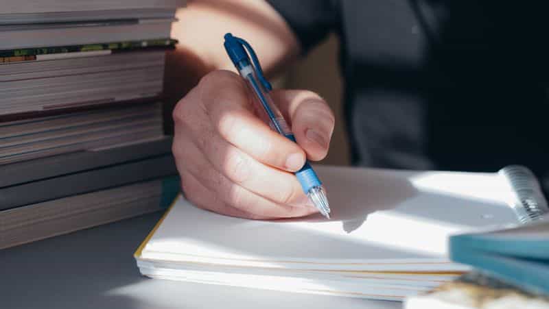 A hand writing with a pen on a pad beside a stack of books in daylight