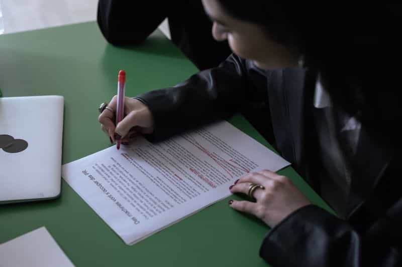 A person marking up a printed page in red pen at a desk beside a closed laptop