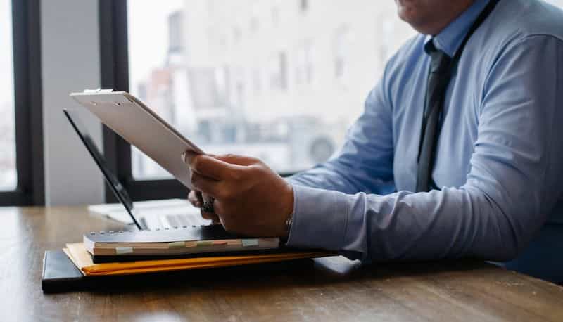 A person in an office reading a document on a clipboard beside a laptop