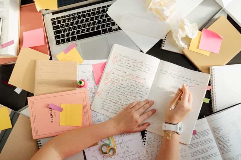 A person writing notes in a notebook at a desk while planning a research paper