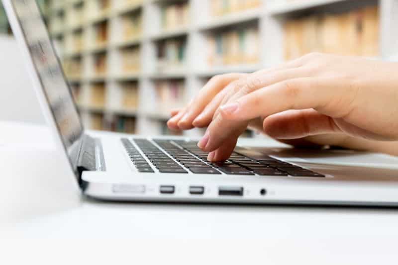 Hands typing on a laptop keyboard in front of library shelves