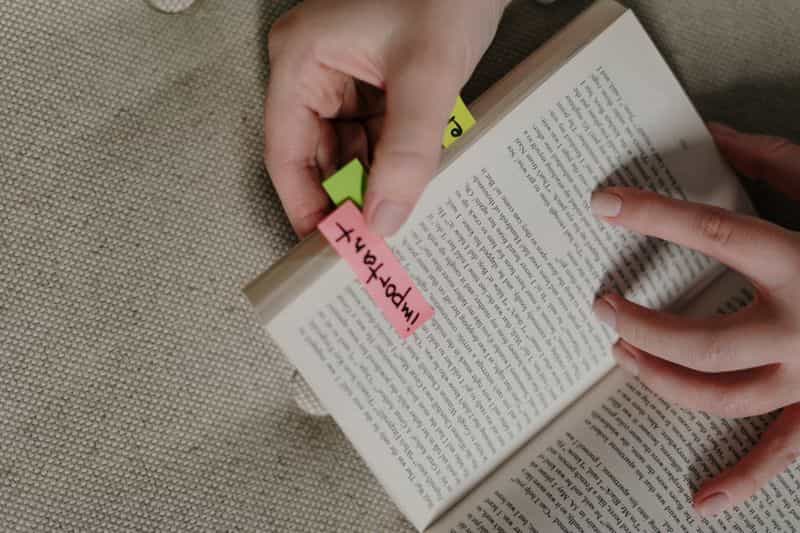 A person holding an open book beside a notebook, taking notes on a source