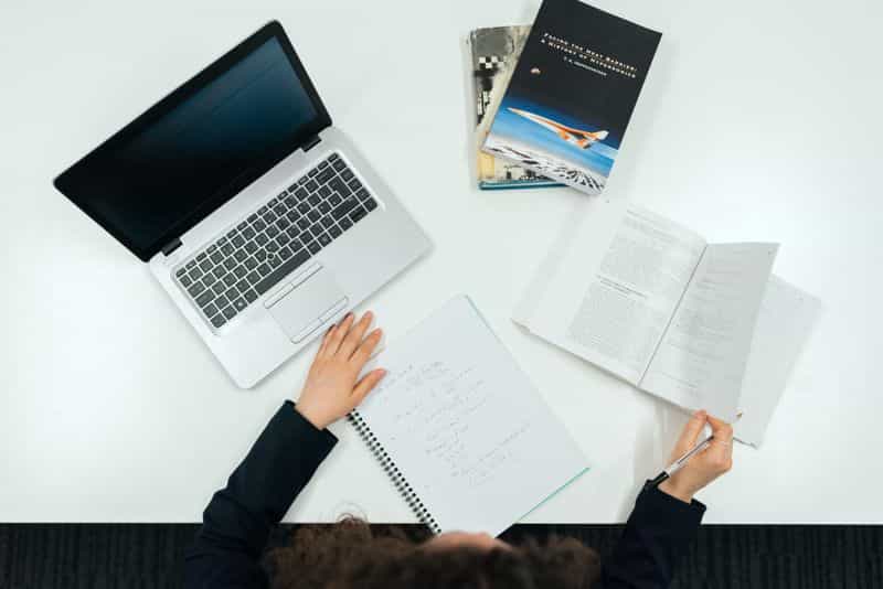 Overhead view of a person at a white desk with an open laptop, a handwritten notebook and an open book, pen in hand