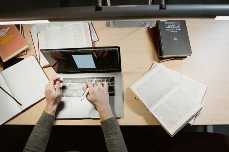 Overhead view of a person typing on a laptop at a library desk, surrounded by open books and a notebook