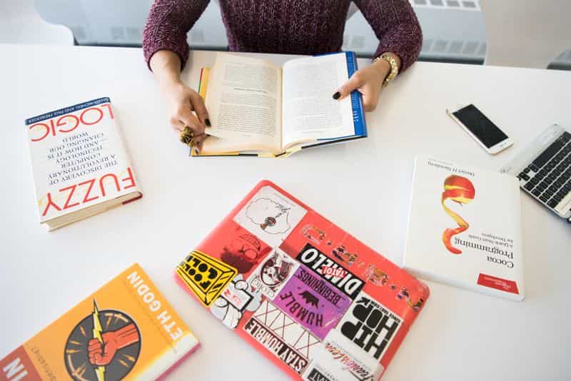 A student's desk with an open book, printed papers, and a laptop showing an organised reading list