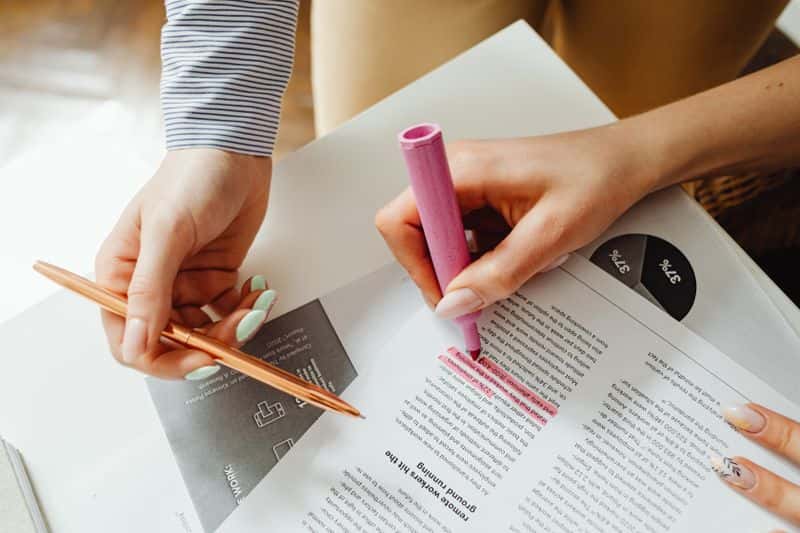 Two hands over printed reports on a desk, one holding a pen and the other marking a passage with a pink highlighter