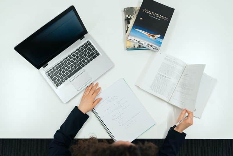 Overhead view of a student at a white desk with an open laptop, handwritten notes in a spiral notebook and an open textbook, pen in hand