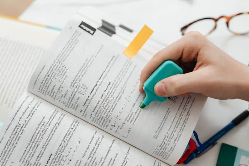 A person marking a line of printed text with a blue highlighter pen, other pages and a notebook on the desk beside them