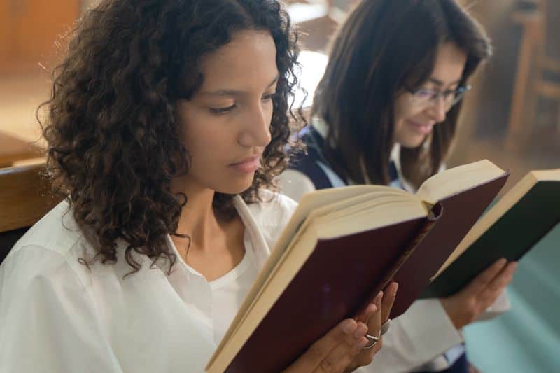Two students reading hardback books side by side at a library table, one book held open in front of the other