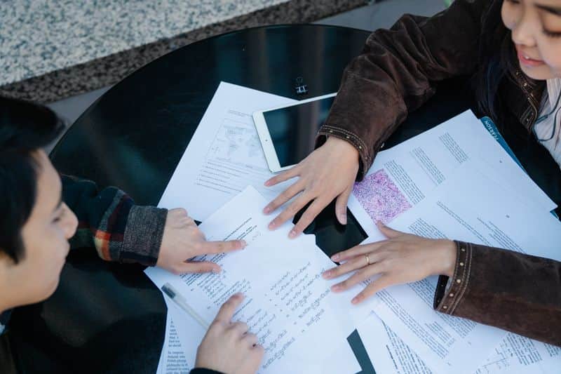 Two students at a round table pointing at printed research papers spread out beside a tablet
