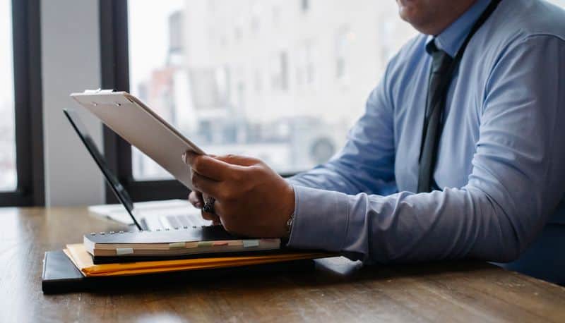 A person in an office reading a document on a clipboard beside a laptop