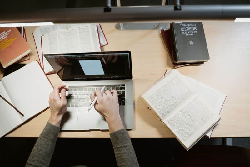 Overhead view of a person typing on a laptop at a library desk, surrounded by open books and a notebook