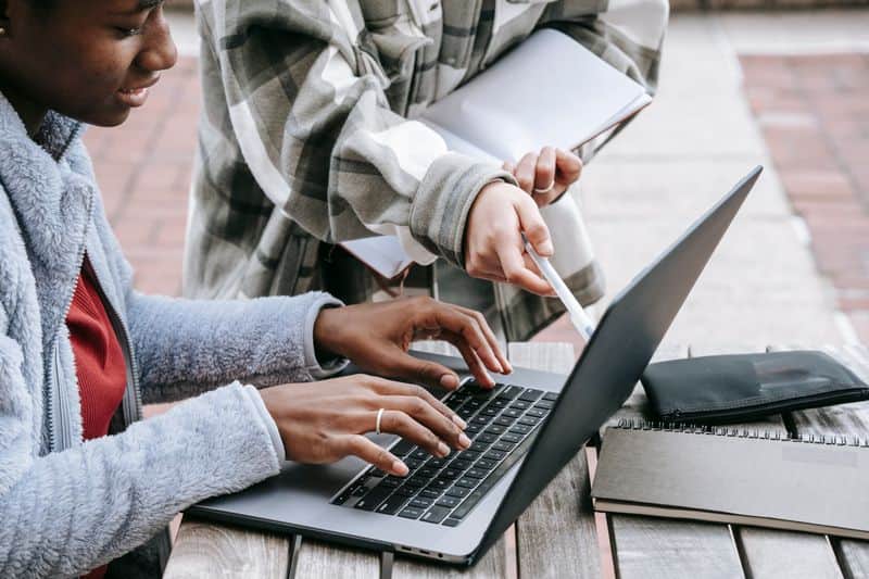 Two students at a laptop on a campus bench, one pointing at the screen while the other types, notebooks beside them