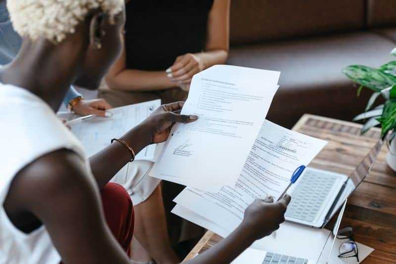 A student reading through printed pages held in one hand, pen in the other, with an open laptop and another person's notes on the table