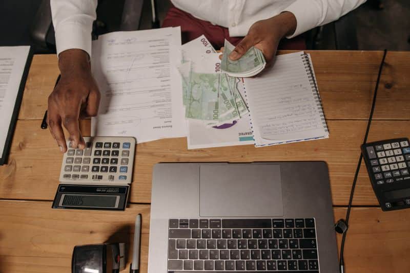 A person sitting at a desk counting banknotes beside a calculator, a notebook and an open laptop