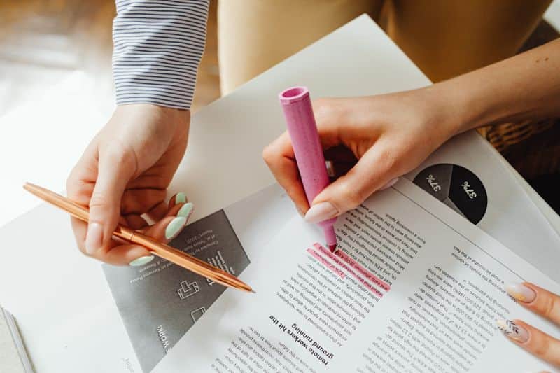 Two hands over printed reports on a desk, one holding a pen and the other marking a passage with a pink highlighter