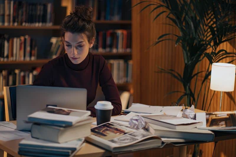 A student working at a laptop in a library, desk covered in stacked books, open journals and a coffee cup