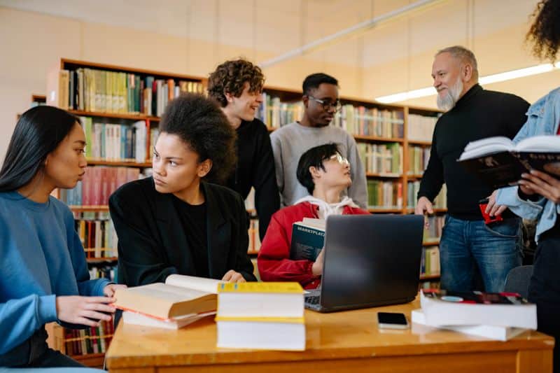 A lecturer talking with a group of students around a laptop and open books at a library table