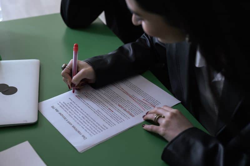 A person marking up a printed page in red pen at a desk beside a closed laptop